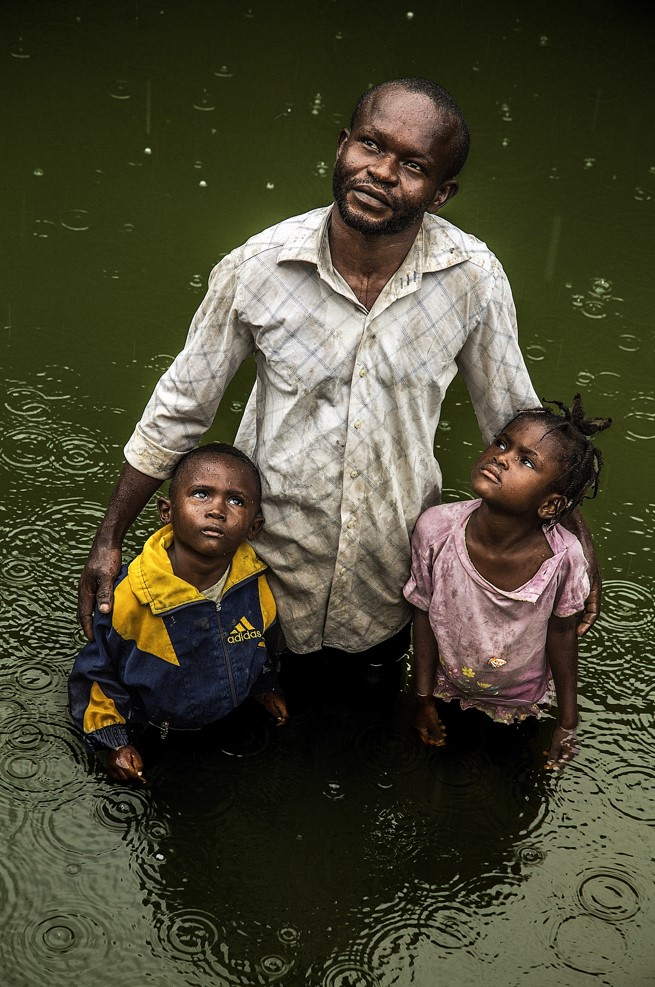 Ciel de saison · Baudouin Mouanda · Moukarata et ses deux enfants observent les nuages dans la cour inondée d'eaux de pluie